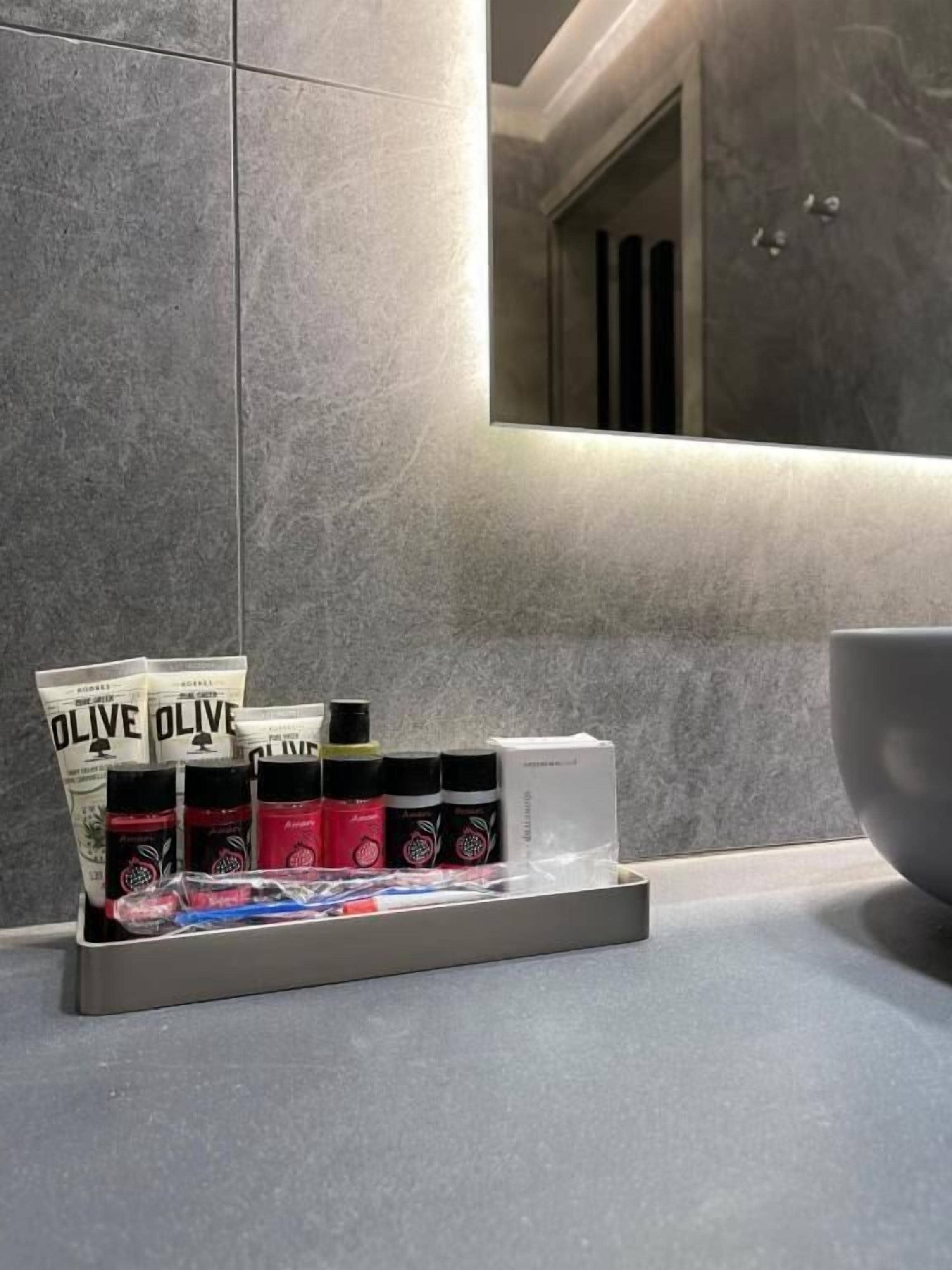 Une salle de bain avec des carreaux gris, un miroir et une étagère contenant divers produits de toilette, notamment de l’huile d’olive et d’autres produits de soins personnels.