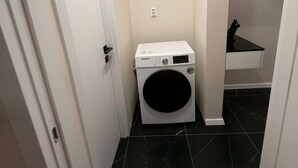 A compact laundry room with a front-loading washing machine, a small countertop, and a door leading to another room. - SteRo Luxury Residence | Modern Apt | Self Check-In | Free Minibar (Galați)