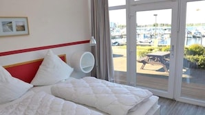 Habitación de hotel con cama, mesita de noche, lámpara y vistas a un puerto deportivo con barcos.