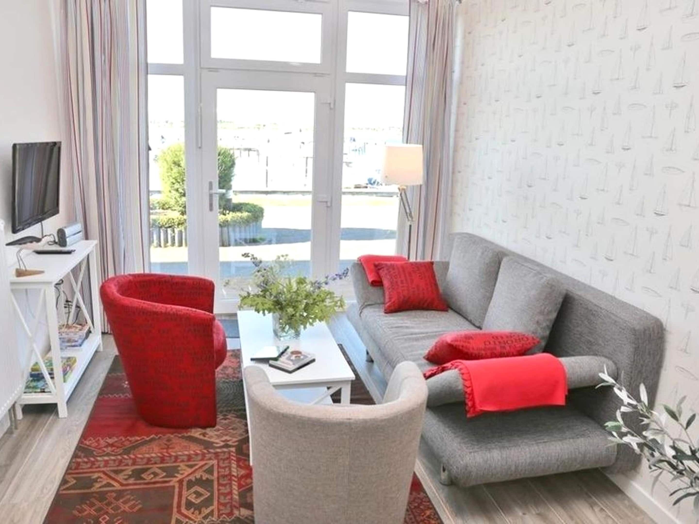 A living room with a grey sofa, red cushions, a red chair, a small table with a plant, and a TV on a white stand.