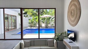Una sala moderna con un ventanal grande que da a una piscina y abundante vegetación.