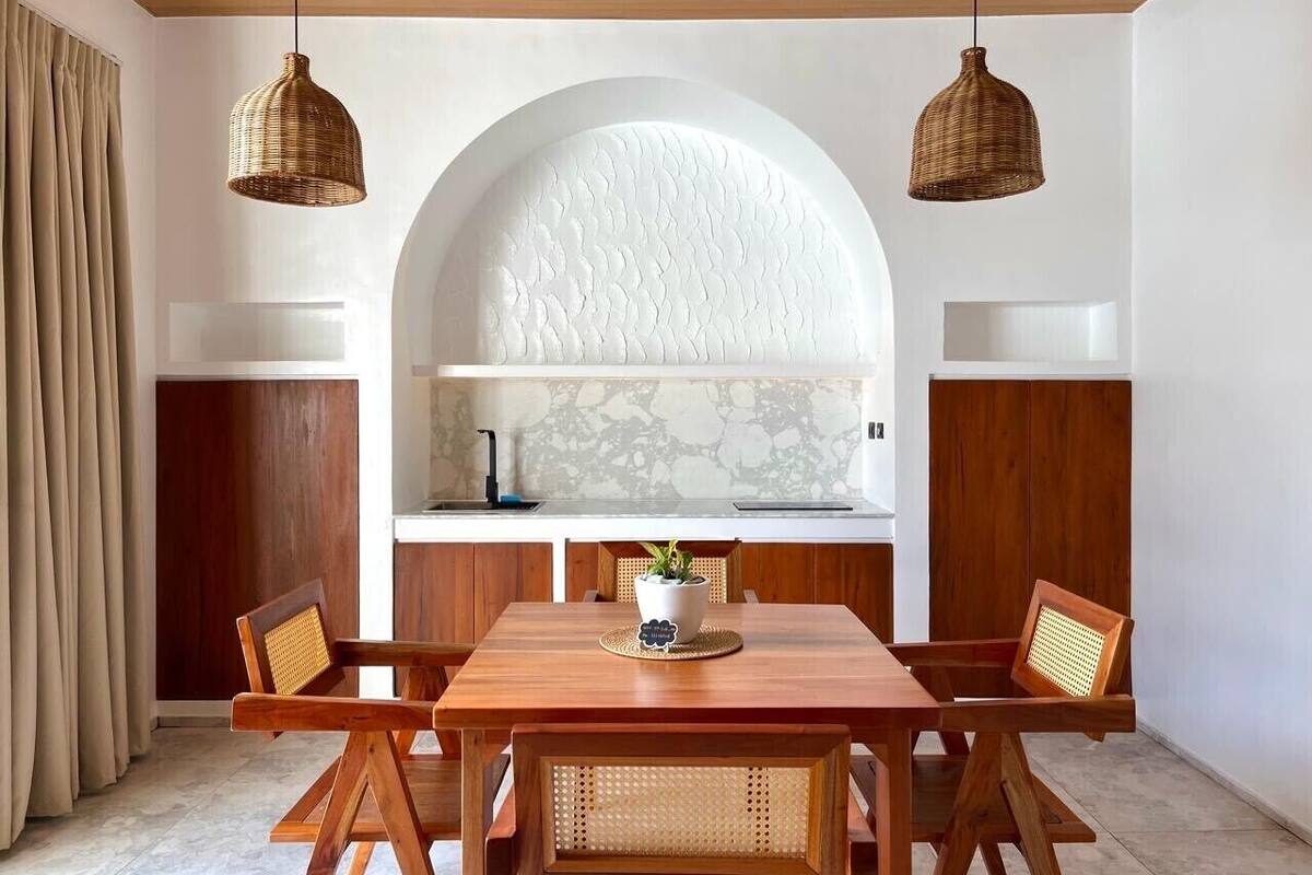 Un coin repas avec une table et des chaises en bois, un mur blanc orné d’un motif texturé et deux suspensions en osier.