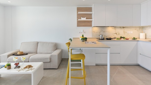 A modern kitchen with white cabinetry, a wooden accent wall, and a dining area with a white table and yellow bar stool. - Cozy Maspalomas Bungalow With Pool (San Bartolomé de Tirajana)