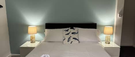 Una cama bien hecha con una colcha blanca y dos almohadas, una manta a rayas y una toalla doblada sobre la cama.