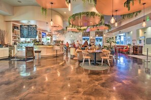 Ein geräumiges Café mit poliertem Betonboden, einer zentralen Sitzgruppe mit runden Tischen und Stühlen sowie hängenden Pflanzen.