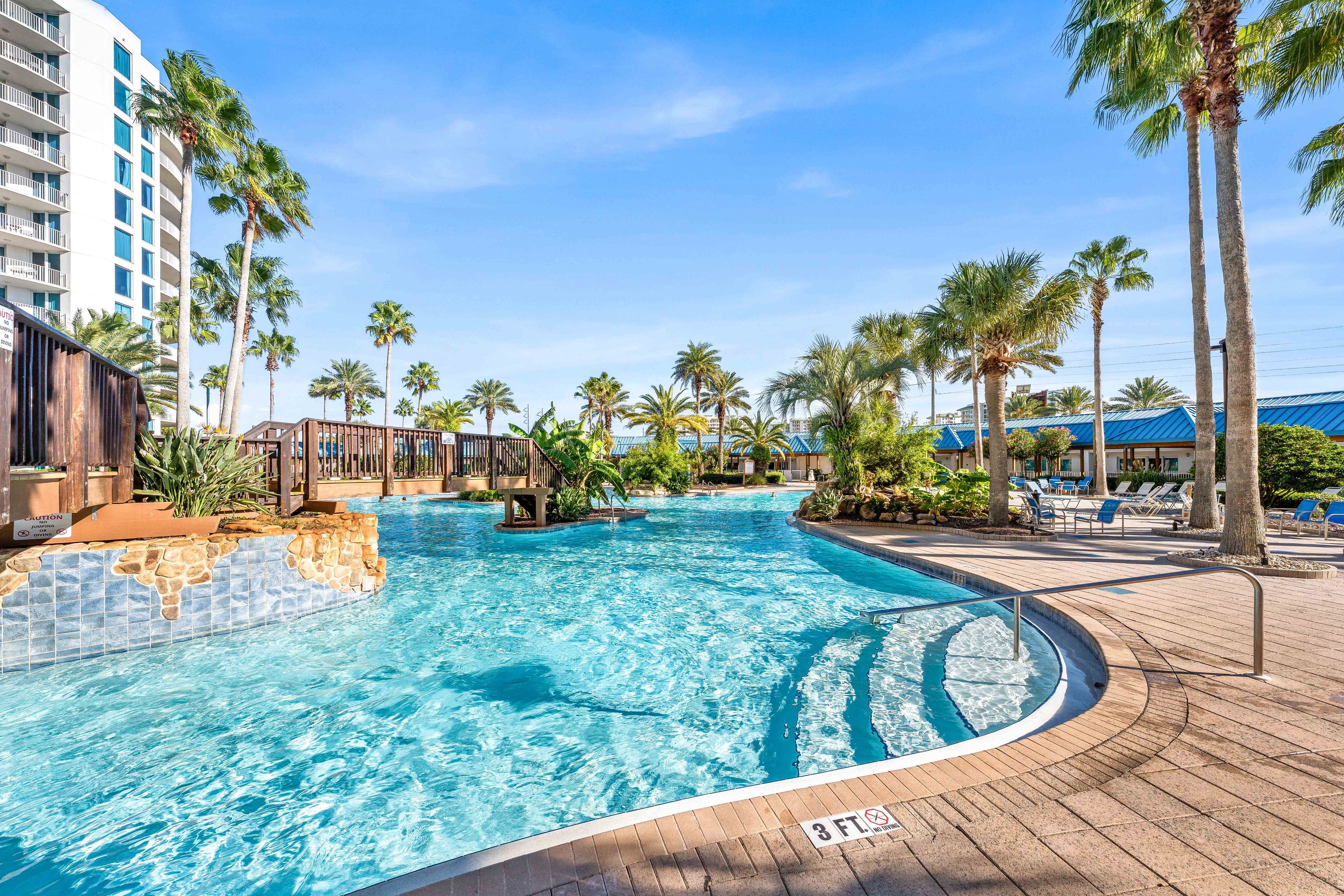 A swimming pool with a wooden deck, palm trees, and a hotel building in the background.
