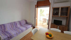 Een kleine kamer met een bank, een tafel met een schaal fruit, een televisie en een schuifdeur naar een buitenruimte.