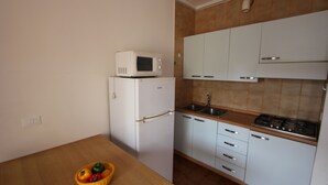 Een compacte keuken met witte kastjes, een magnetron, een spoelbak en een fornuis. Er staat een houten tafel met een mand vol fruit.