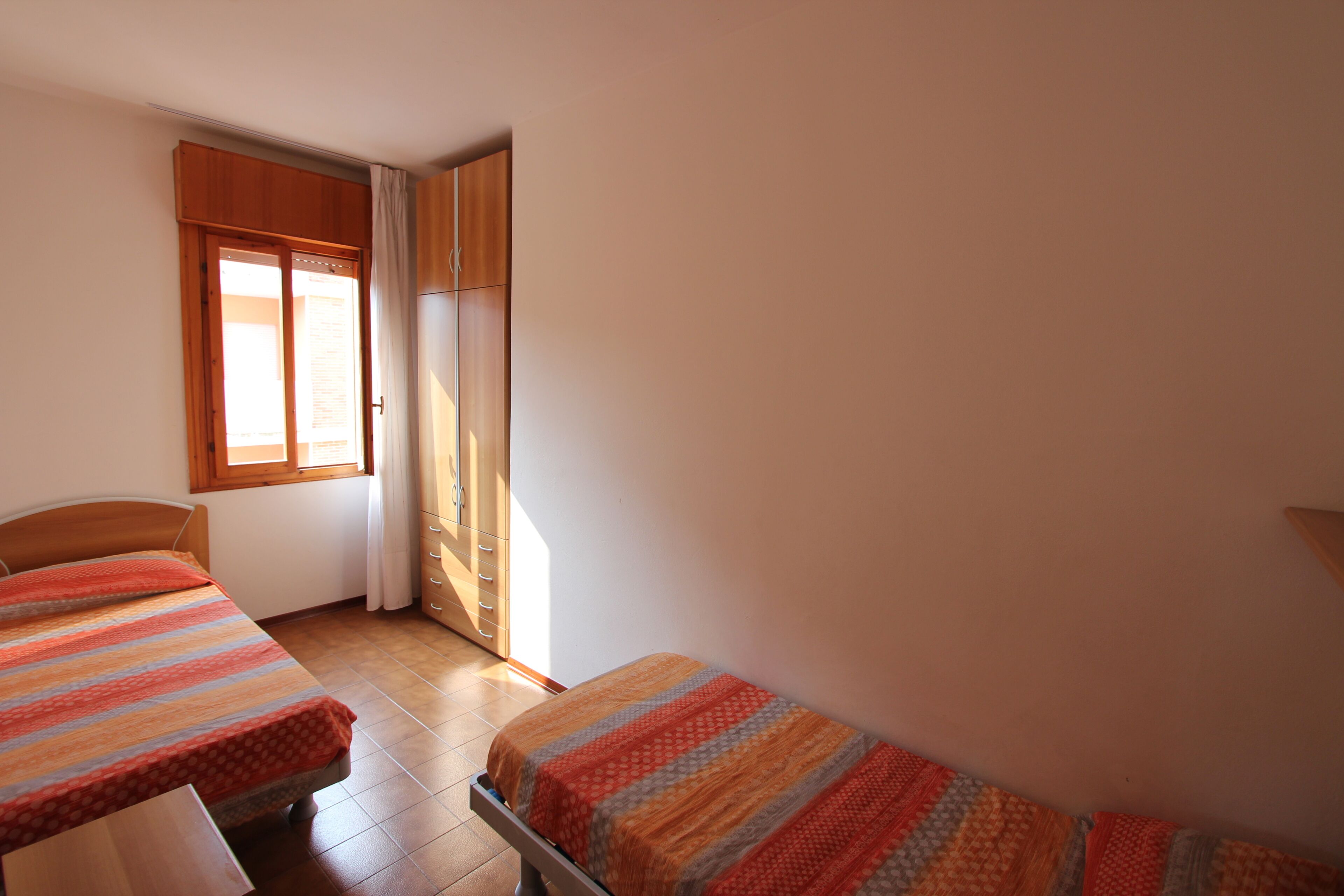 A bedroom with two beds, a wooden wardrobe, a window with curtains, and a tiled floor.
