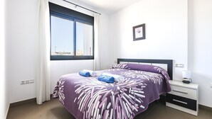 Een net opgemaakt bed met een bedsprei met patroon, een nachtkastje met een lamp, en een raam met gordijnen.