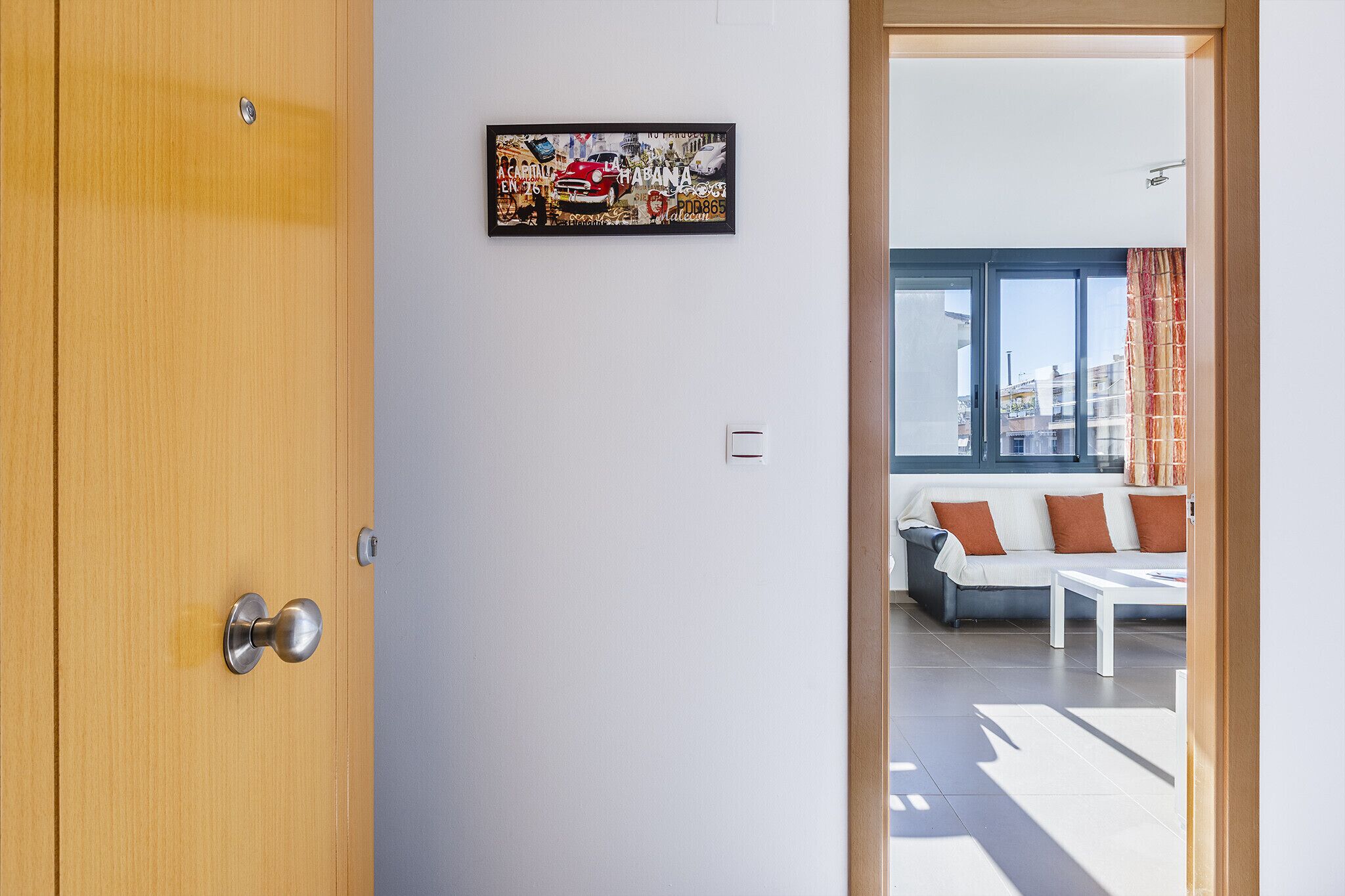 A modern room with a wooden door, a framed artwork on the wall, and a view of a living area with a sofa and a table.