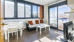 Een moderne woonkamer met een bank, salontafel en televisie, grote ramen met gordijnen, en een balkon met tuinmeubilair.