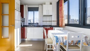 Een compacte keuken met witte kasten, een eettafel met een tafelkleed met bladmotieven, en een raam met uitzicht op gebouwen.