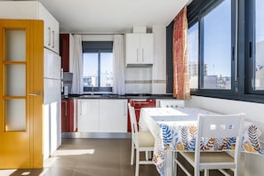 A compact kitchen with white cabinets, a dining table with a leaf-patterned tablecloth, and a window with a view of buildings. - ATICO EDIFICIO ARAGÜEZ (Torre del Mar)