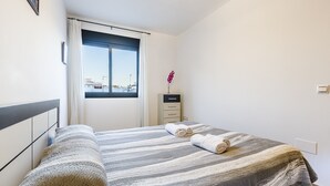 Ein ordentlich bezogenes Bett mit Streifenmuster, ein Fenster mit Vorhängen, ein kleiner Nachttisch mit einer Vase und ein an der Wand befestigter Lichtschalter.