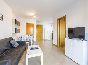 Una sala de estar moderna con un sofá, una mesa de centro, un televisor en un mueble, y un área de cocina al fondo.