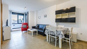 Ein modernes Wohnzimmer mit einem schwarzen Ledersofa, einem roten Sessel, einem Esstisch mit Stühlen, einem Fernseher und einem großen Fenster mit Vorhängen.