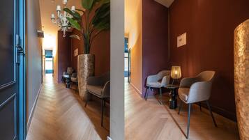Een hotelgangen met houten vloer, een grote potplant en twee zithoekjes met tafels en stoelen.