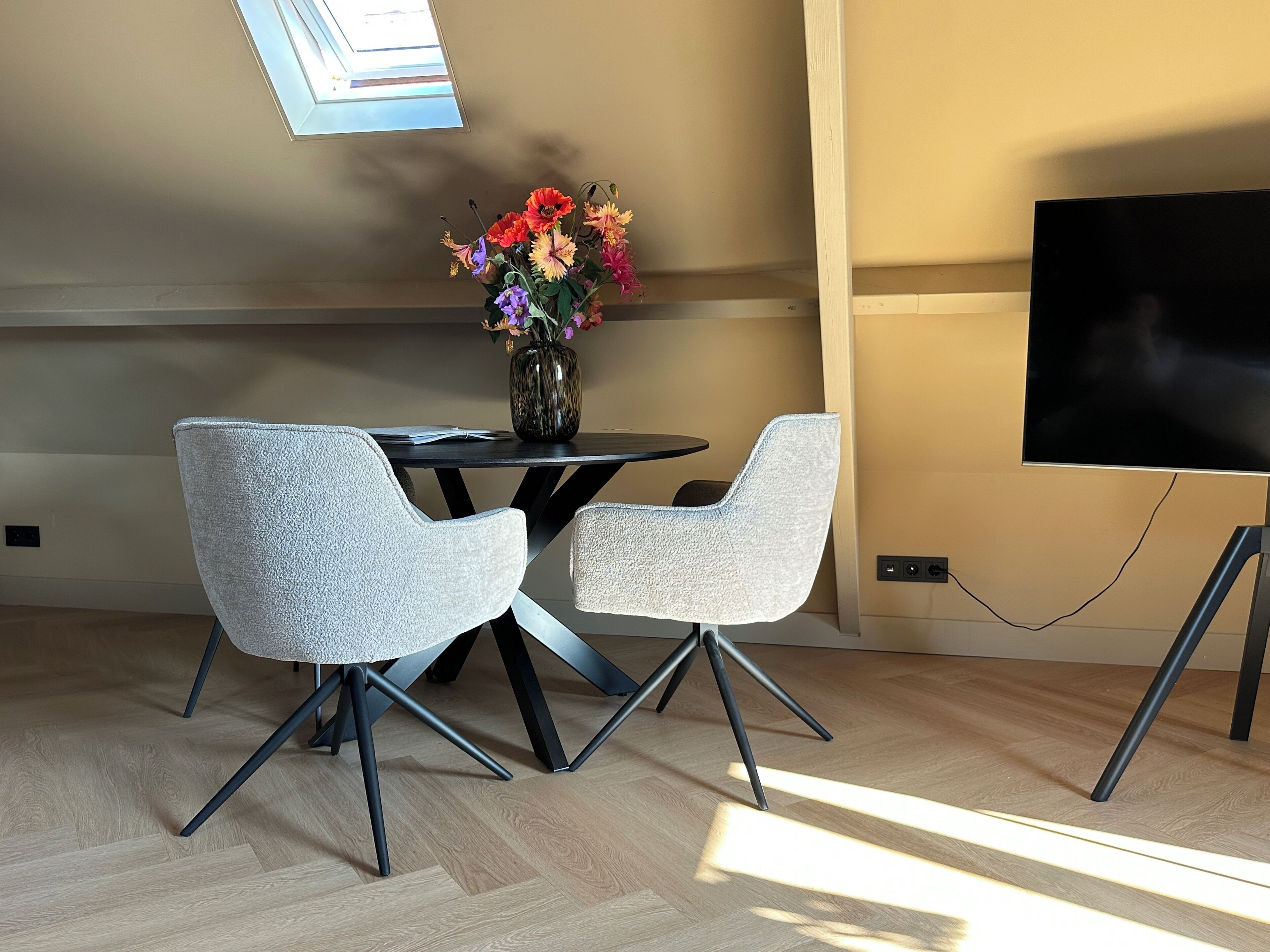 A modern dining area with two grey chairs, a round table, and a vase with colorful flowers. A flat-screen TV is mounted on the wall to the right.