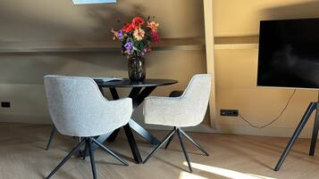 A modern dining area with two grey chairs, a round table, and a vase with colorful flowers. A flat-screen TV is mounted on the wall to the right.