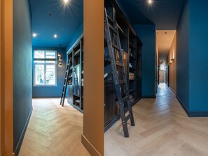 Un couloir au plancher en bois, une échelle appuyée contre un meuble de couleur sombre et une fenêtre avec des stores.