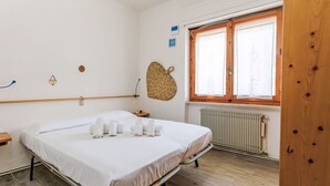 A clean, well-lit room with a bed, a window with wooden frames, a radiator, and a woven wall decoration. - Oleandro by Interhome (Castiglione della Pescaia)