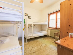 Een slaapzaal met stapelbedden, een houten vloer, een raam met gordijnen, een radiator en een klein tafeltje met een doos erop.