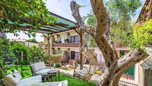 A tree with a swing chair, a covered seating area, and a building with balconies in the background. - Oleandro by Interhome (Castiglione della Pescaia)