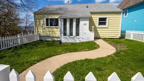 A two-story house with a white picket fence, a driveway, and a clear blue sky. - Adorable Hilltop Cottage by the Beach (Michigan City)
