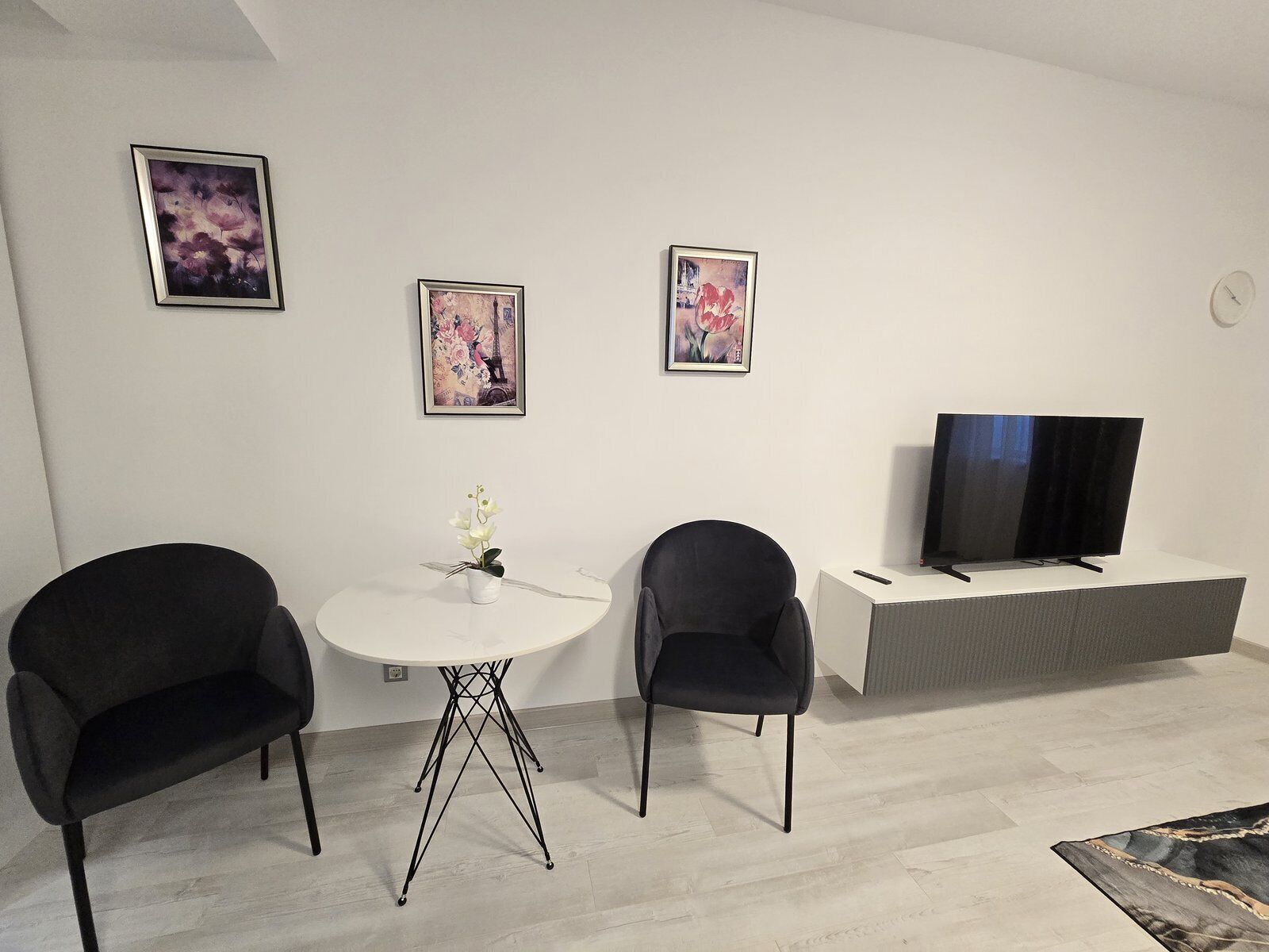 A modern living room with a white round table, two black chairs, a flat-screen TV on a grey stand, and three framed artworks on the wall.