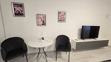 A modern living room with a white round table, two black chairs, a flat-screen TV on a grey stand, and three framed artworks on the wall.