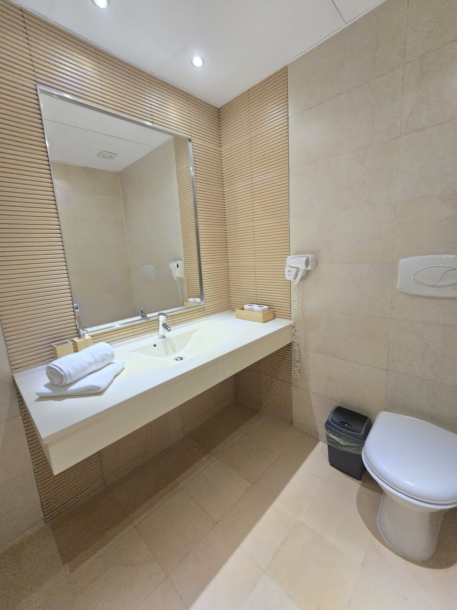 A modern bathroom with a long sink, a mirror, a toilet, and a trash bin.