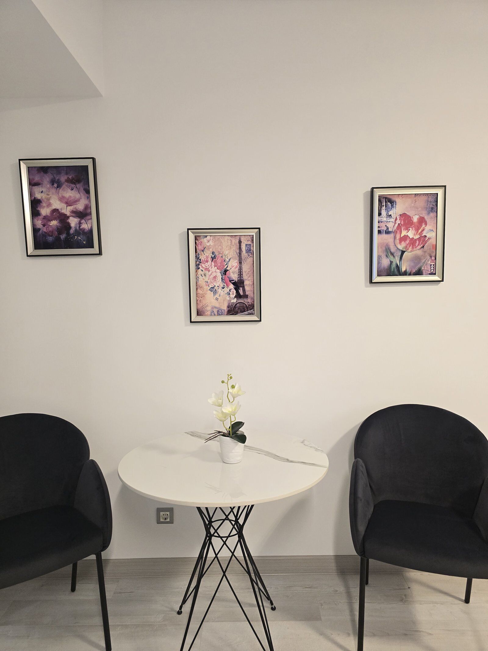 Una habitación minimalista con una mesa redonda, dos sillas negras y tres obras de arte enmarcadas en la pared.