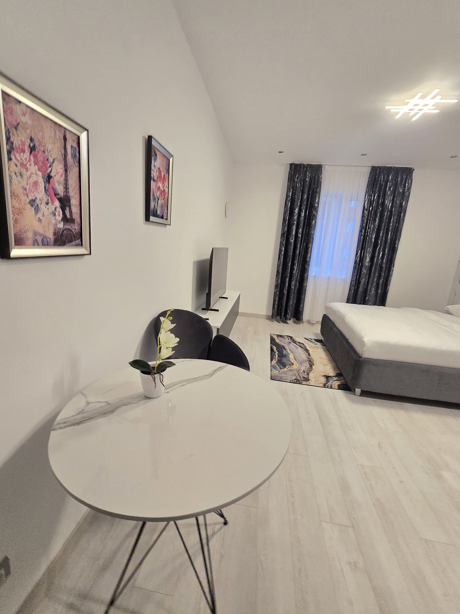 A modern hotel room with a round dining table, two black chairs, a bed, a large window with curtains, and two framed artworks on the wall.