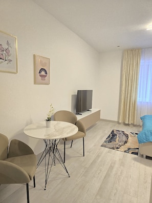 A modern living room with a round dining table, beige chairs, a flat-screen TV on a low shelf, a blue cushion on a white sofa, and a patterned rug on the floor.