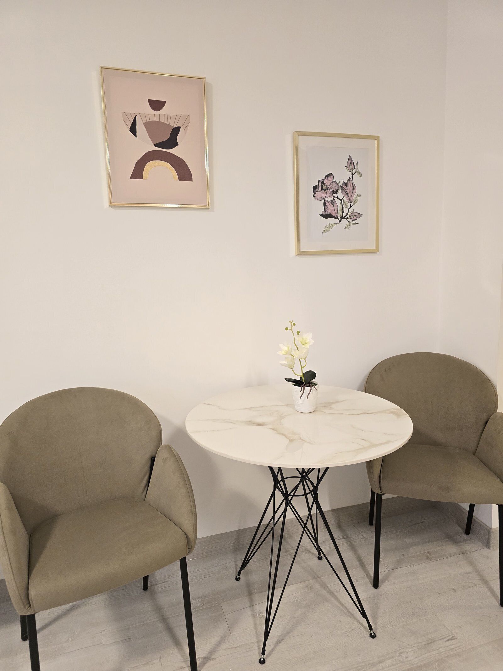 A room with a round marble table, two beige chairs, and two framed artworks on the wall.