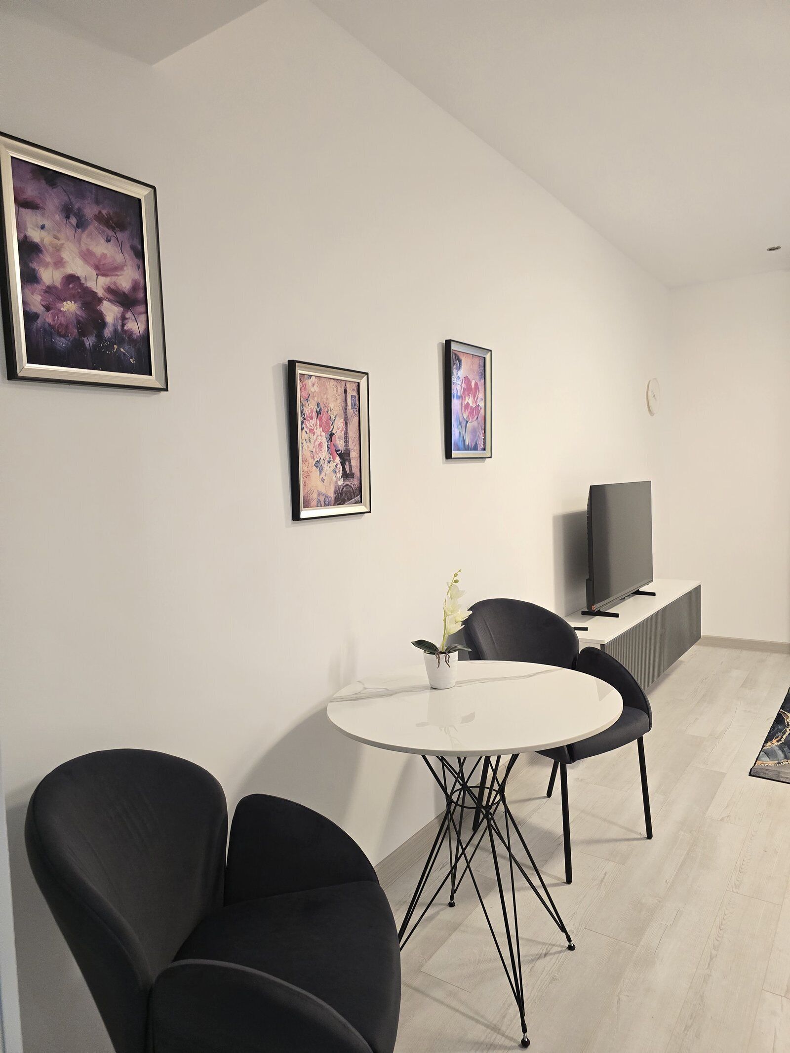 Un salon moderne avec une table ronde blanche, deux chaises noires, une plante en pot blanche, un téléviseur à écran plat et deux œuvres d’art encadrées accrochées au mur.