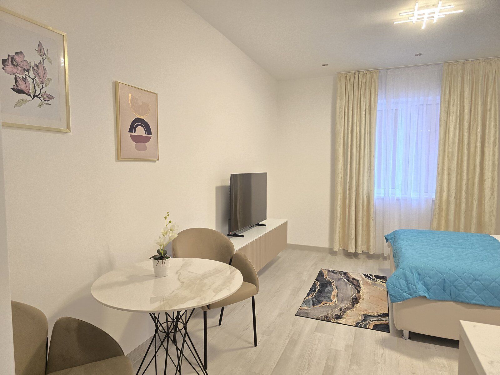 A modern hotel room with a bed, a TV, a dining table with chairs, and a small potted plant.