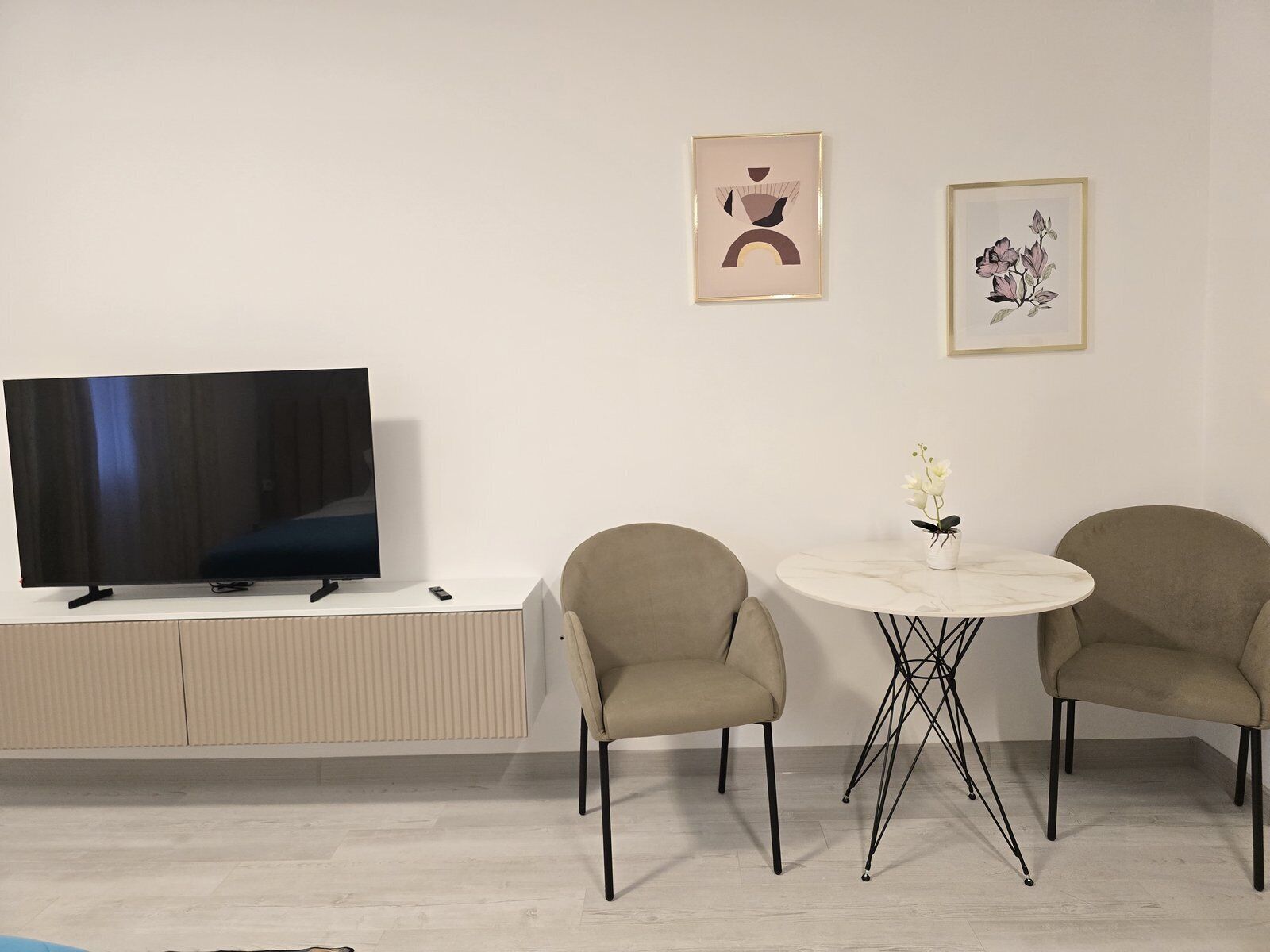 Ein modernes Wohnzimmer mit einem Flachbildfernseher auf einem weißen Schrank, zwei beigefarbenen Sesseln um einen kleinen runden Tisch mit einer Blumenvase und zwei gerahmten Kunstwerken an der Wand.
