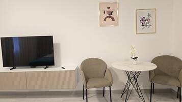 Ein modernes Wohnzimmer mit einem Flachbildfernseher auf einem weißen Schrank, zwei beigefarbenen Sesseln um einen kleinen runden Tisch mit einer Blumenvase und zwei gerahmten Kunstwerken an der Wand.