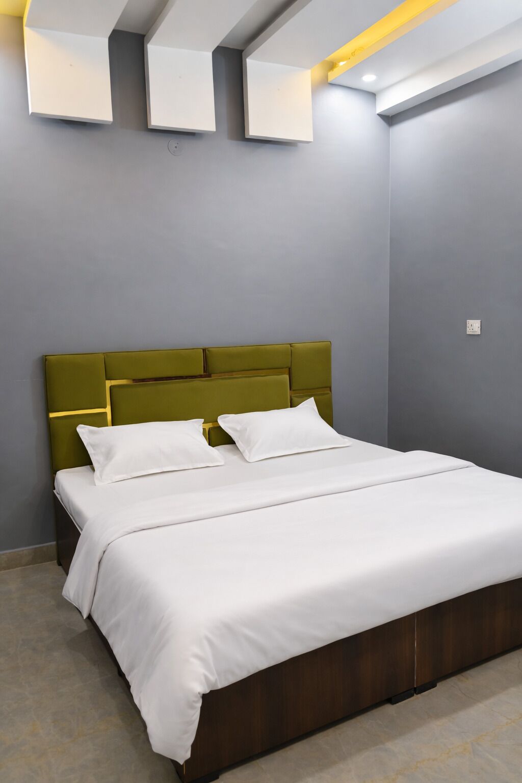 A neatly made bed with white bedding and a green headboard in a modern room with grey walls and recessed lighting.
