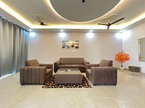 A modern living room with a sofa set, a coffee table, a giraffe-patterned rug, ceiling fans, recessed lighting, and a wall-mounted painting. - Book N Go HomeStay - Greater Noida (Greater Noida)
