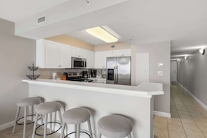 A modern kitchen with white cabinets, a stainless steel refrigerator, a microwave, and a bar area with stools. - Gulf front home with shared pool, private balcony and beach access (Fort Walton Beach)