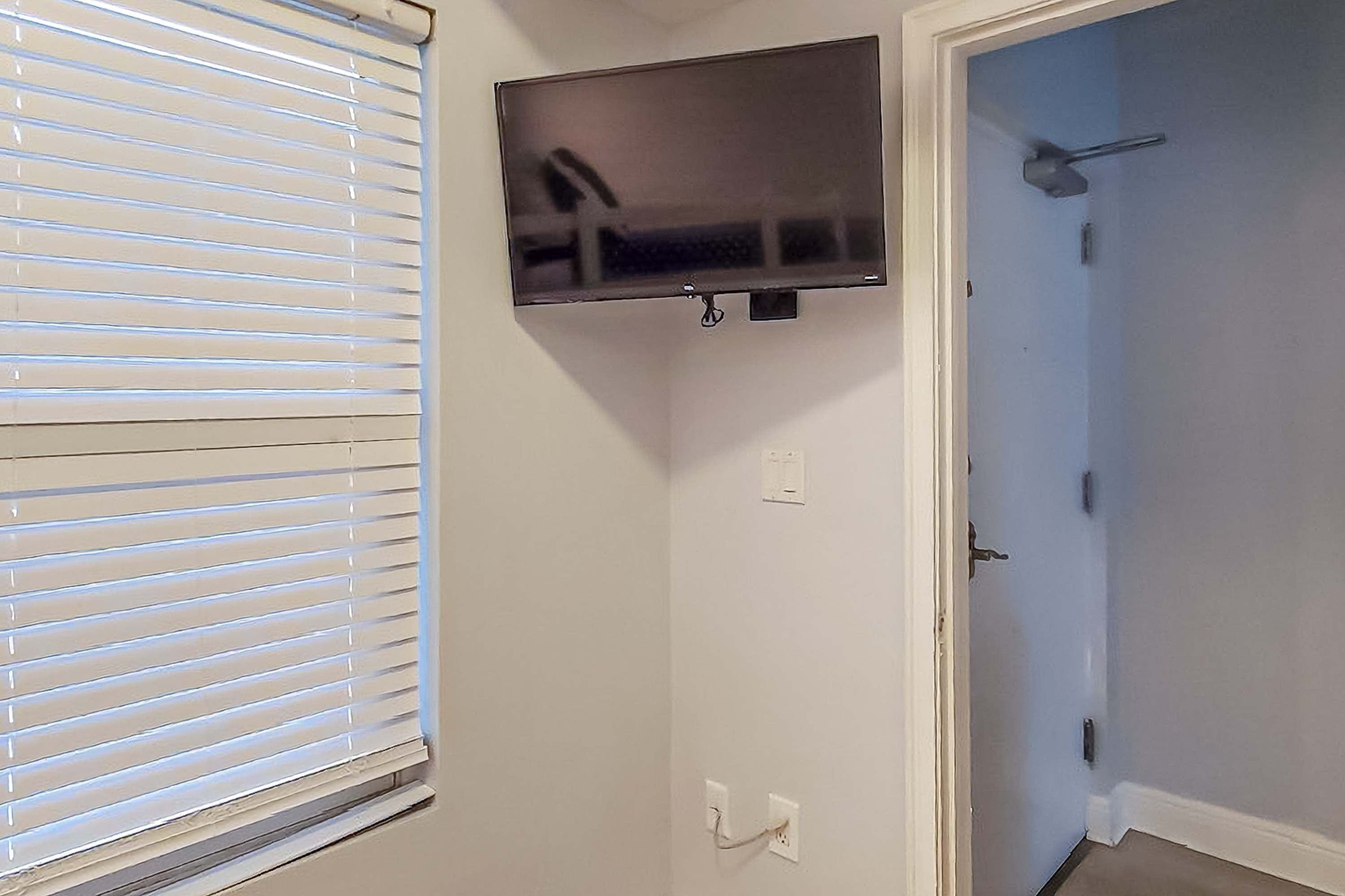 Una habitación con una televisión instalada en la pared, una puerta con manija y una ventana con persianas.