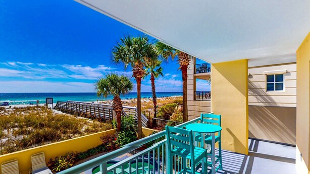 Ein Balkon mit türkisfarbenen Stühlen und einem Tisch, Blick auf einen Strand mit Palmen und einem strahlend blauen Himmel.