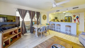 Ein modernes Wohnzimmer mit Fernseher, Esstisch und Stühlen, einer Küche mit Barhocker und einem Deckenventilator.