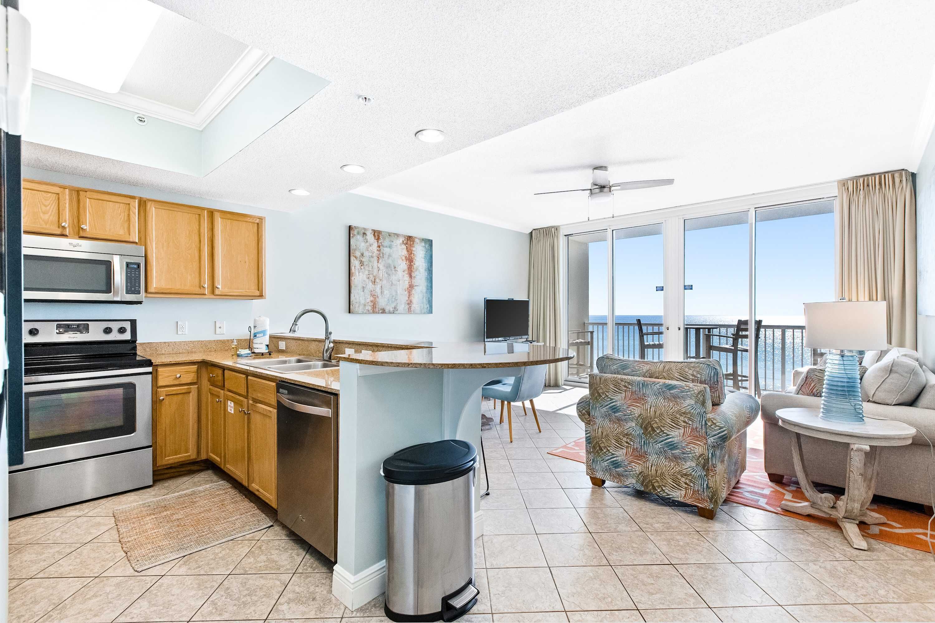 A modern kitchen with a microwave, oven, and sink. There is a dining area with a chair and a table. The room has a large window and a ceiling fan.