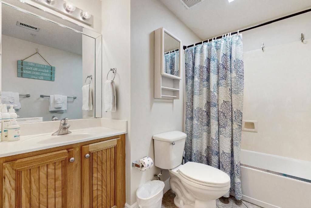 Une salle de bain avec un meuble-lavabo en bois, des toilettes, un rideau de douche orné d’un motif d’arbre et un miroir au-dessus du meuble-lavabo.