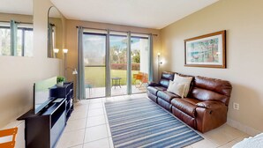A living room with a brown leather sofa, a flat-screen TV, and a large window with curtains. - Serene studio with garden-level patio, central AC, waterfront pool & marina (Fort Walton Beach)