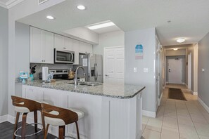 A kitchen with white cabinets, a granite countertop, stainless steel appliances, and two wooden bar stools. - Gulf Front condo with heated pool, beach access, gym, washer&dryer and Wifi (Fort Walton Beach)